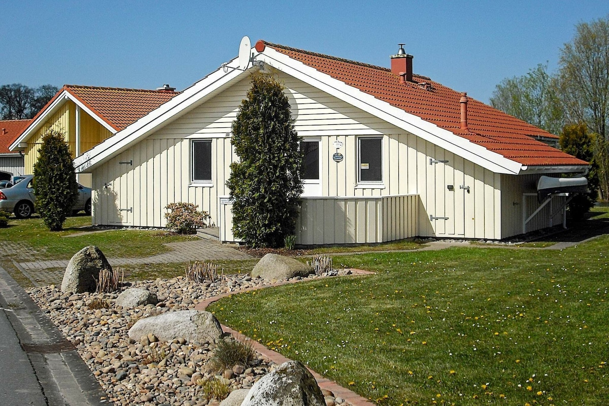 The exterior of a holiday home is depicted, showcasing a well-maintained yellow and white structure with a red-tiled roof. Surrounding greenery includes manicured lawns and a decorative rock garden, enhancing the inviting appearance of the property. The home features large windows and a chimney.
