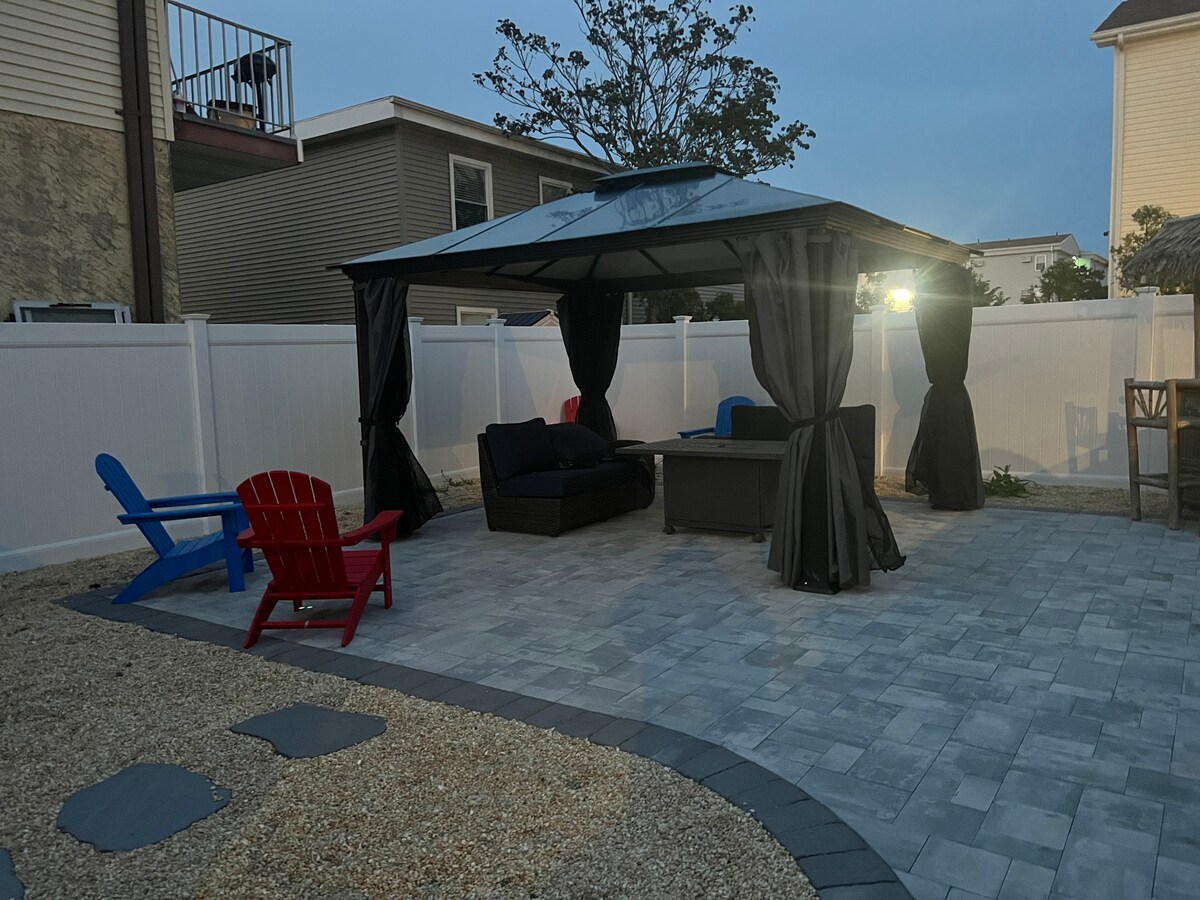 A shaded outdoor seating area is framed by a gazebo with flowy drapes. Comfortable seating is arranged around a central table, with red and blue chairs positioned nearby. The patio is made of stone pavers and surrounded by a neat gravel landscape.