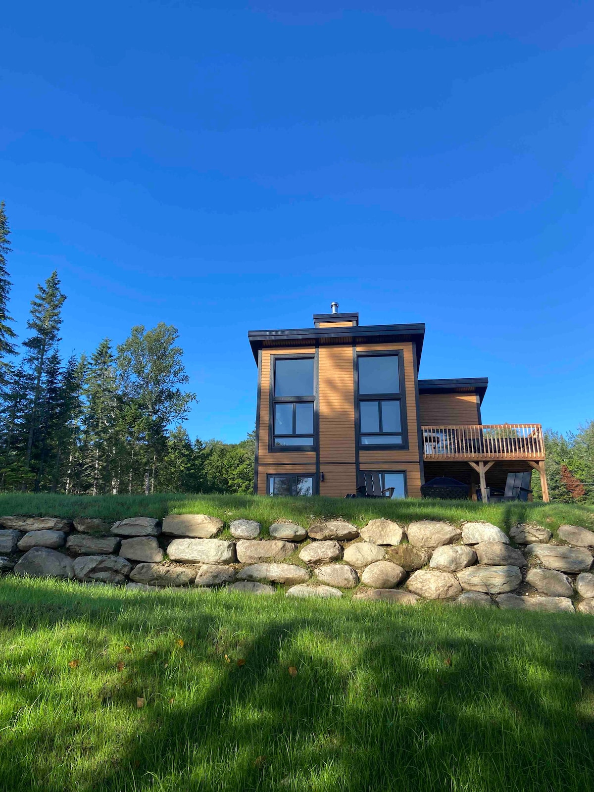A modern chalet structure is seen from a lower angle, showcasing large windows and a balcony. The surrounding landscape features lush green grass and a rock wall, complemented by tall trees in the background under a clear blue sky.