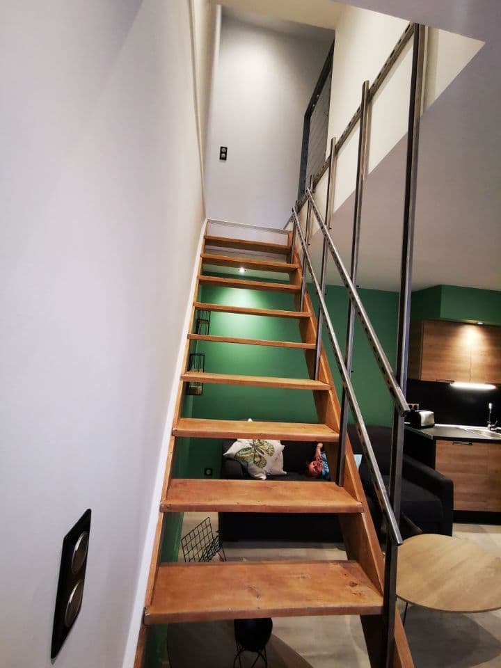 A wooden staircase is shown leading to an upper level. The walls are painted in a soft color, while modern lighting highlights the space. A glimpse of a green wall and contemporary kitchen area is visible in the background, suggesting a cohesive design throughout the apartment.