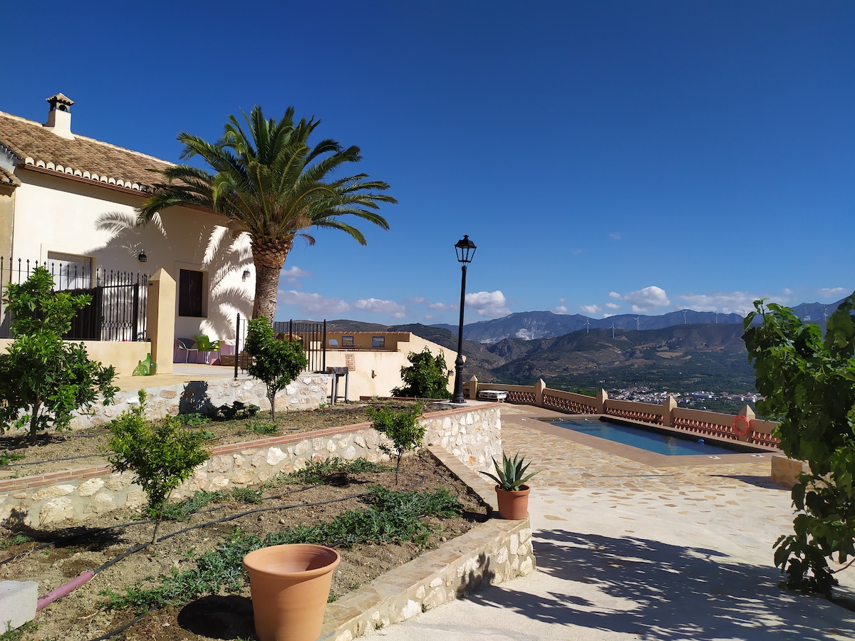 A charming building is situated among landscaped gardens, featuring palm trees and a variety of plants. The property offers sweeping views of the surrounding mountains and valley, with a stone pathway leading to a tranquil pool area in the background.