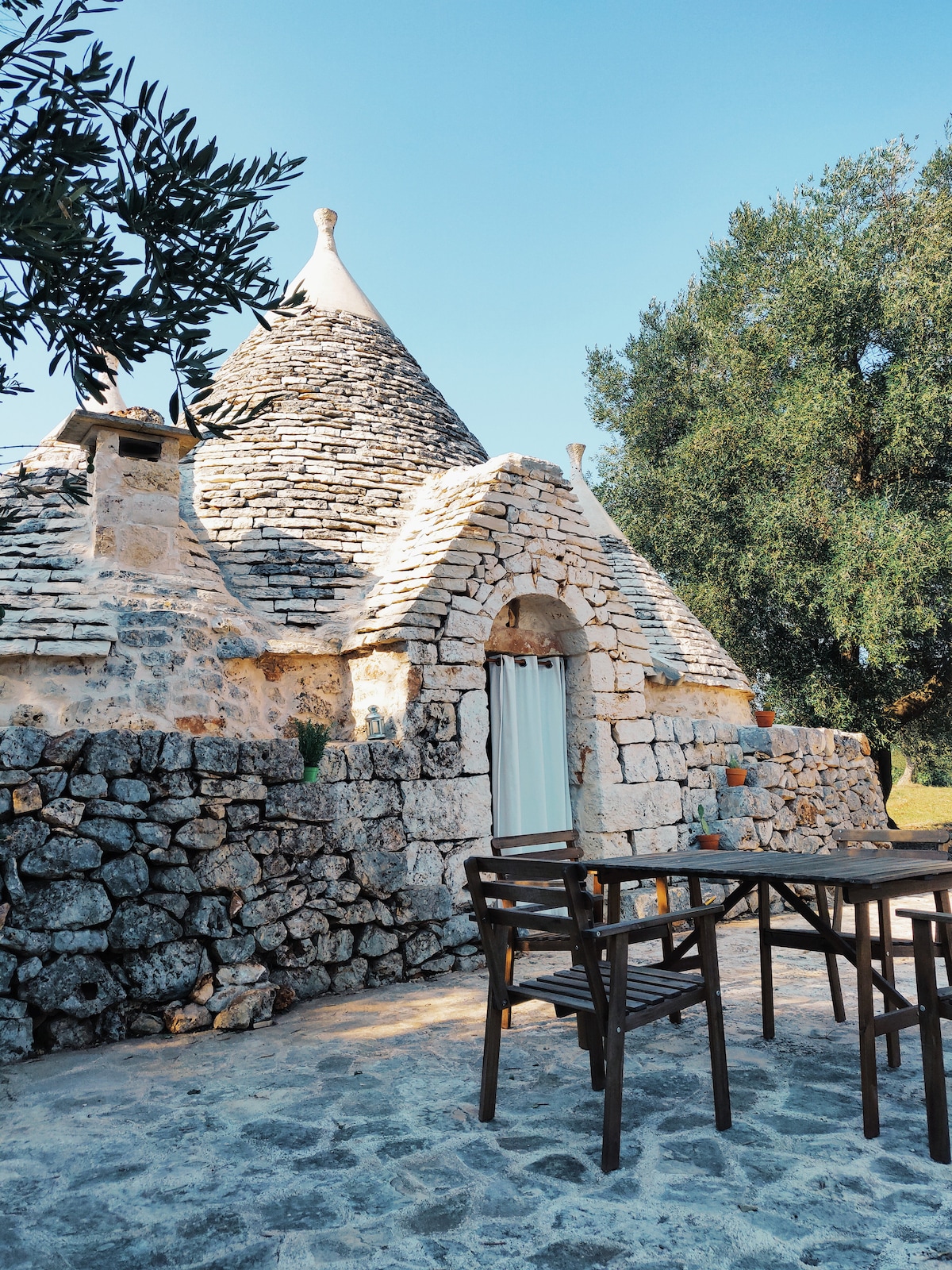 The traditional trullo structure is captured under a clear blue sky, featuring a stone facade and a conical roof. A wooden table and chairs are positioned on a stone patio, surrounded by lush olive trees, inviting a relaxed outdoor experience.