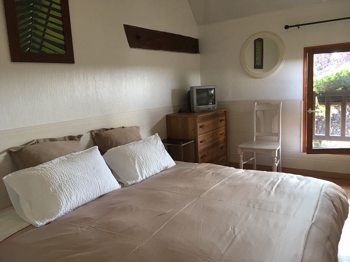 A spacious sleeping area is furnished with a large bed topped with neutral-colored linens and two pillows. A wooden dresser and a white chair accompany the room, and a small television is placed on the dresser. Natural light enters through a round window overlooking greenery.