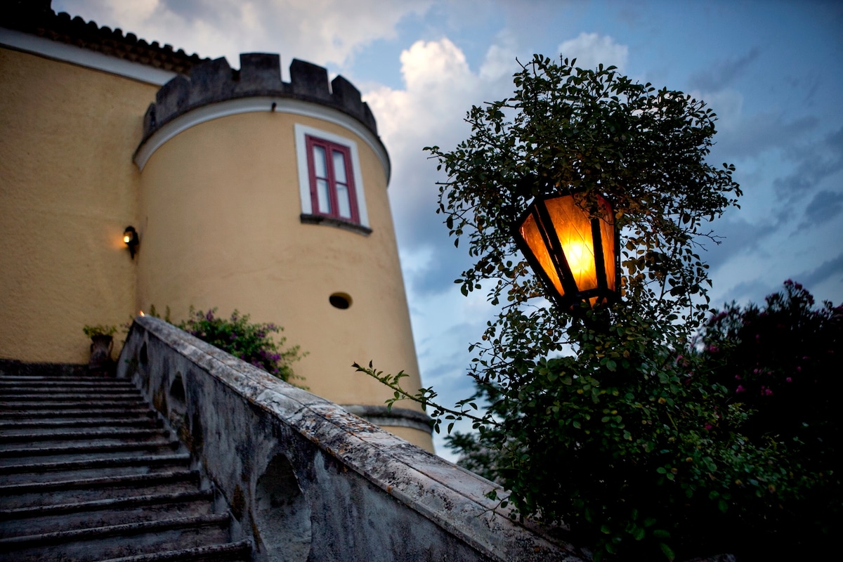 A stone staircase leads up to a tower-like structure highlighted by warm lantern light. The building features a rounded wall with a turret and red window frames. Lush greenery surrounds the staircase, enhancing the outdoor ambiance as twilight descends.