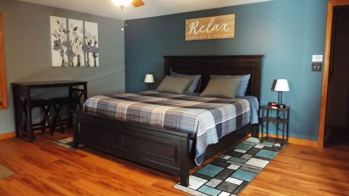 A spacious bedroom is furnished with a king bed dressed in plaid bedding. A small dining table is positioned nearby, accompanied by two light-colored table lamps. Wall art featuring floral themes decorates the space, while rich wood flooring adds warmth.