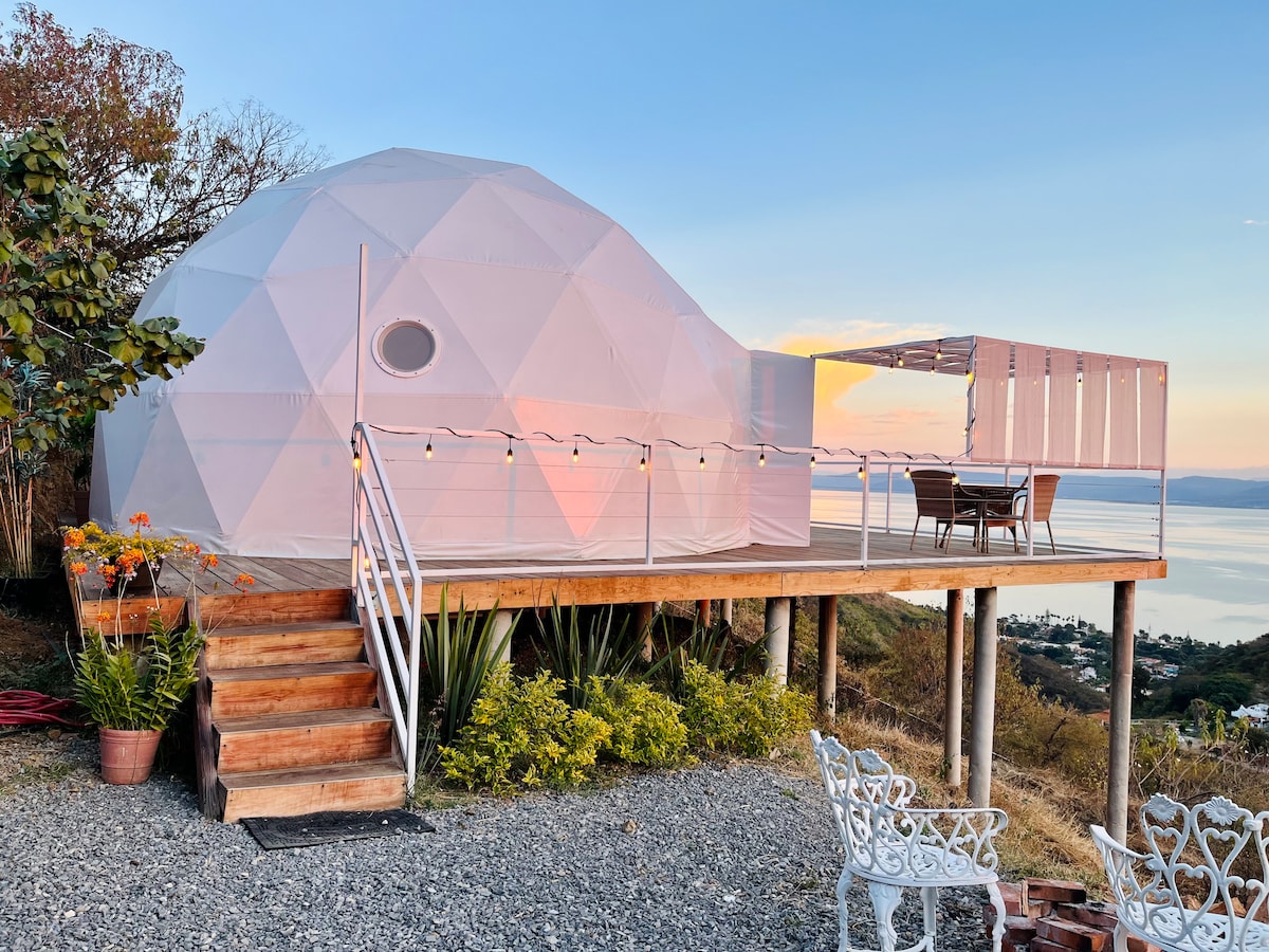 A geodesic dome structure is elevated on a wooden platform, surrounded by natural landscaping. A spacious terrace extends from the dome, featuring a dining set and outdoor seating. Soft, warm lighting accents the upper edges of the dome, while panoramic views of the lake and mountains are visible.