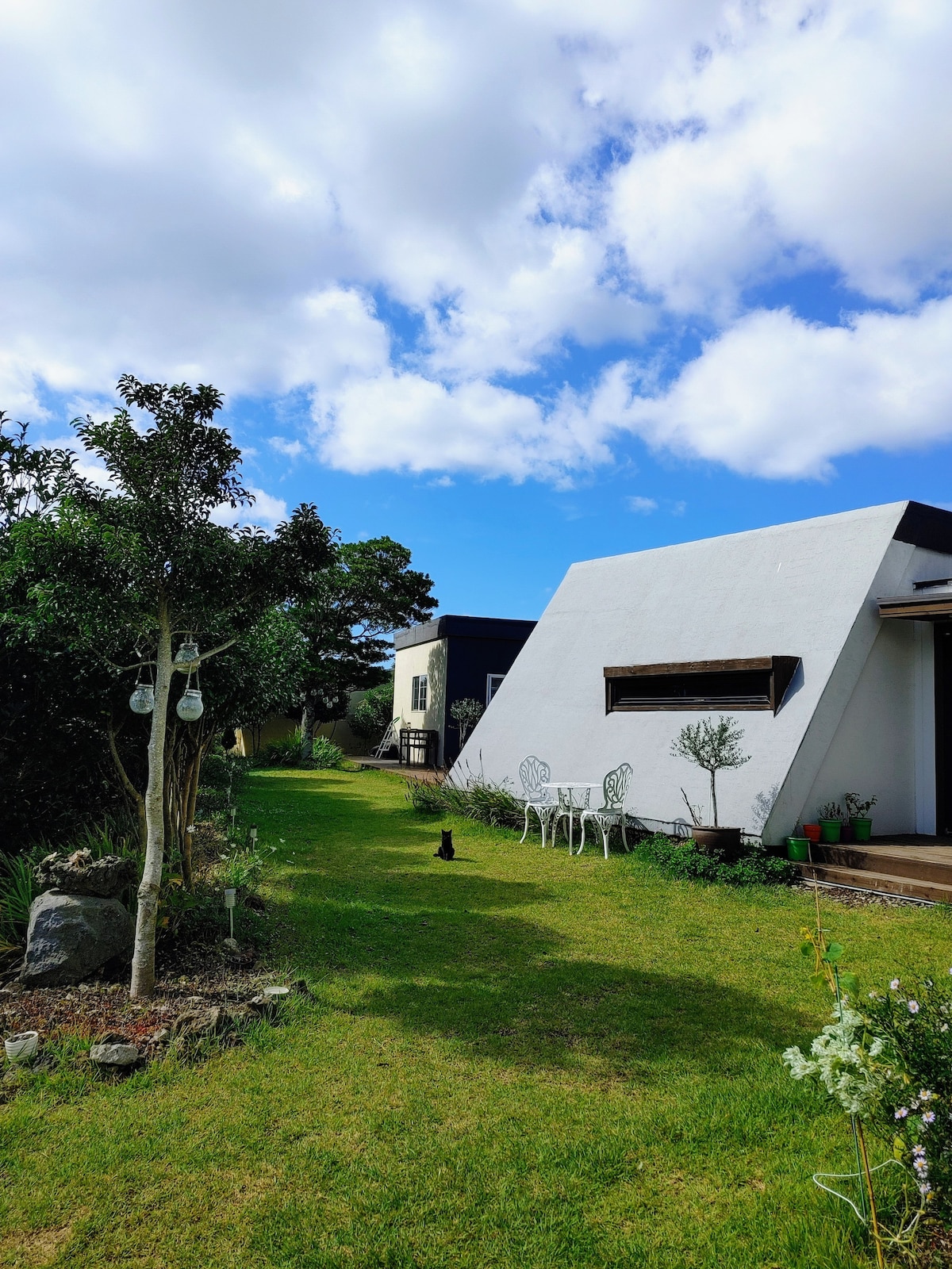 A well-maintained grassy pathway leads to a modern white building with an angled roof. Two white chairs are positioned near the entrance, surrounded by greenery and small plants. The blue sky and fluffy clouds create a serene backdrop.