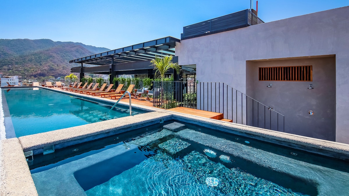 A rooftop area features a sleek infinity pool with a gradual edge, reflecting the clear blue sky. Beside it, a smaller hot tub offers a relaxing option. The sun deck is adorned with comfortable lounge chairs and greenery, providing an inviting space with mountain views.
