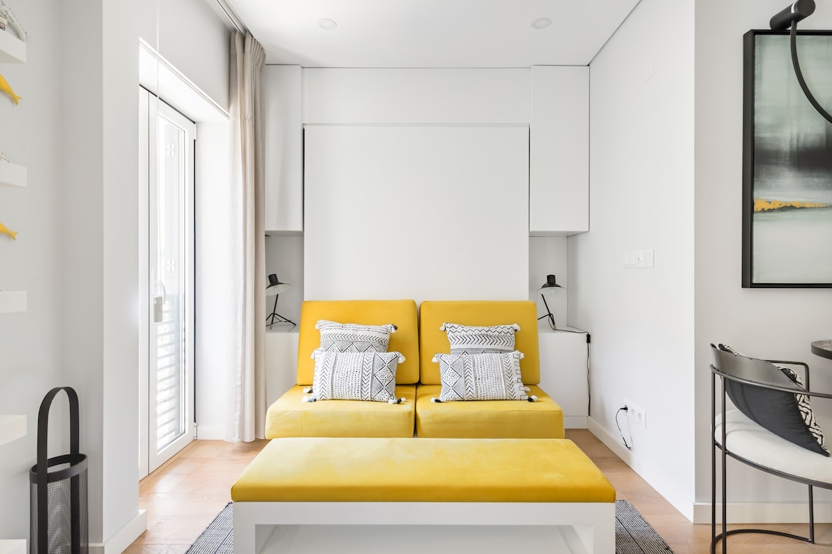 A contemporary yellow sofa is positioned in a bright living area, featuring two decorative pillows. A minimalist coffee table sits in front, while a large painting adorns the wall. Natural light enters through the balcony door, enhancing the airy feel of the space.