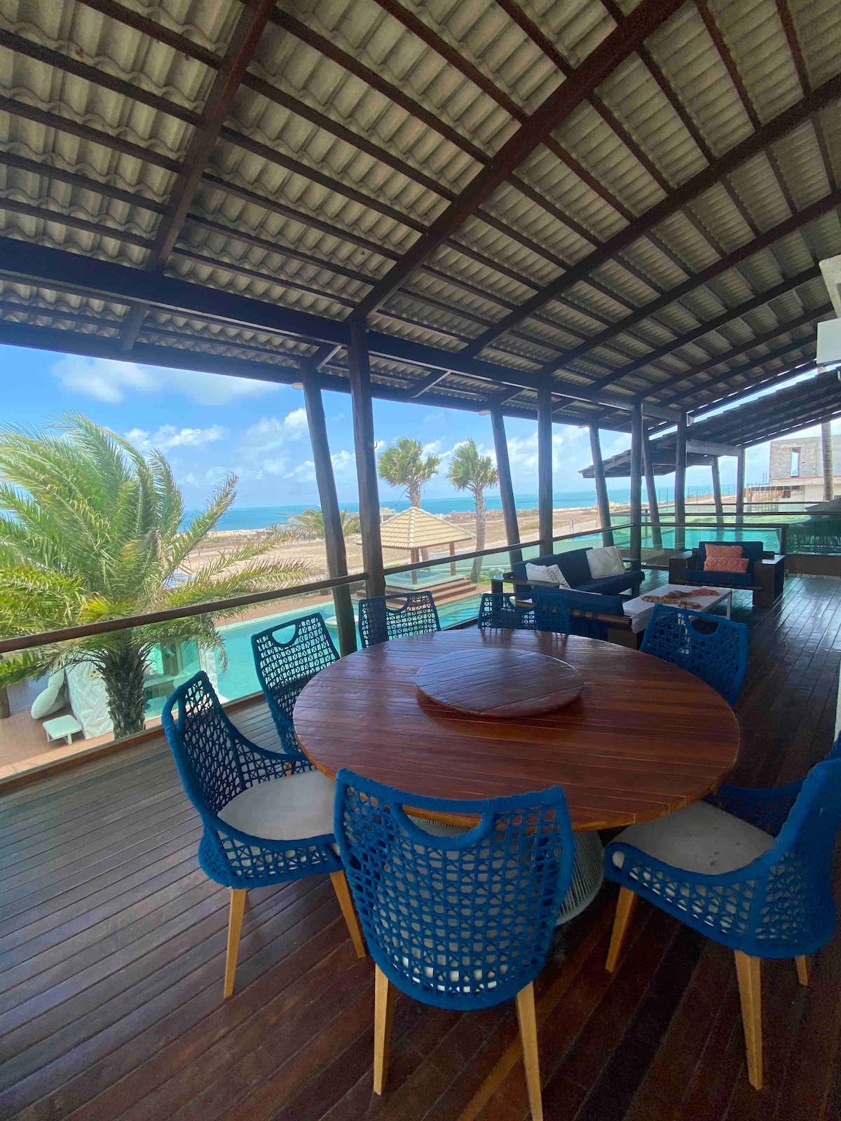 An outdoor dining area is visible, featuring a large round wooden table surrounded by six blue woven chairs. A seating arrangement with cushions is present, and palm trees can be seen swaying outside, with a glimpse of water in the background under a clear sky.