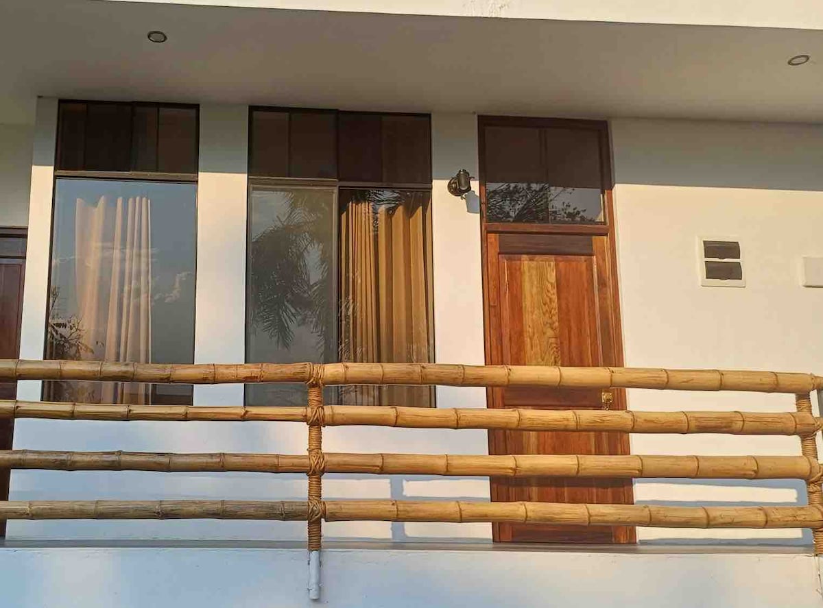 The exterior of the building features a simple wooden railing and a door crafted from natural wood, complemented by large windows with sheer curtains allowing natural light to filter inside. The entrance is accentuated by a softly lit wall sconce.
