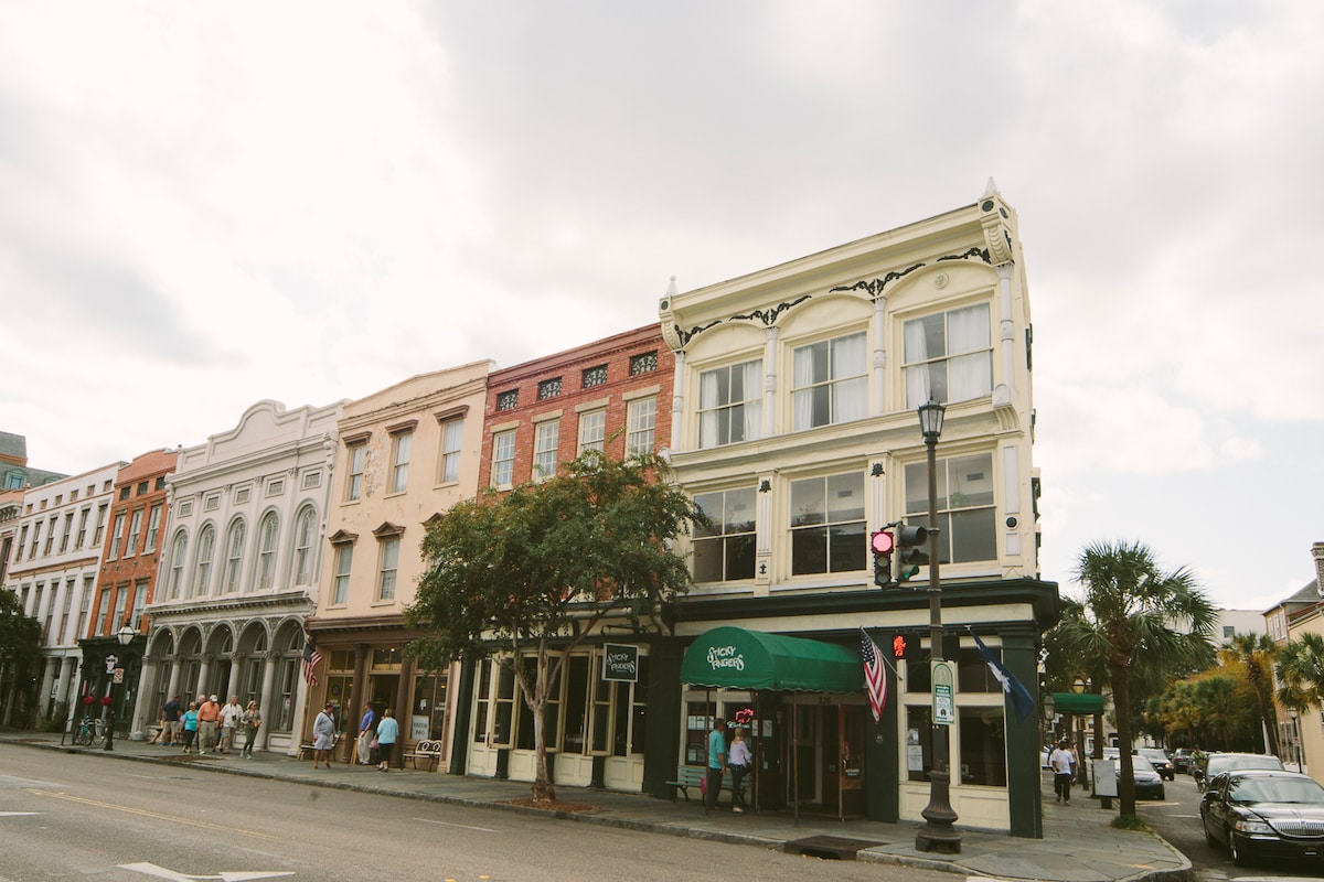 A charming street view presents a row of historic buildings with varied architectural styles, characterized by vibrant façades. Palm trees are visible alongside the sidewalk, while pedestrians enjoy the lively atmosphere. A streetlight stands in the foreground, completing the urban scene.