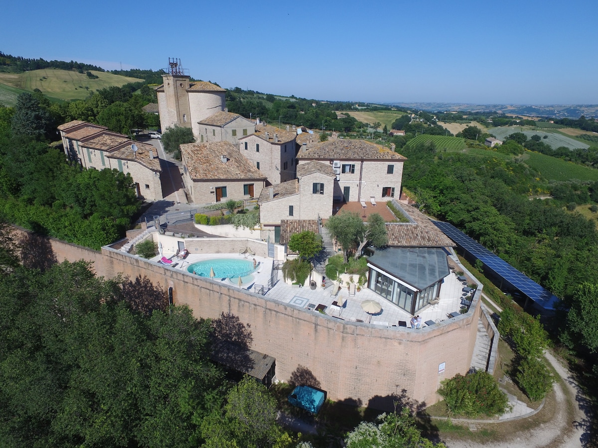 Aerial view of a multi-structure property set amidst lush greenery, featuring a swimming pool surrounded by lounge chairs. The landscape includes rolling hills, olive trees, and picturesque buildings, showcasing a harmonious blend of nature and architecture.