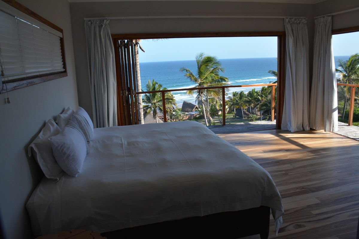 A light-filled bedroom features a spacious bed with white linens. Large glass doors open to a balcony, providing views of the ocean and surrounding palm trees. Natural wood flooring adds warmth to the space, enhancing the coastal ambiance.