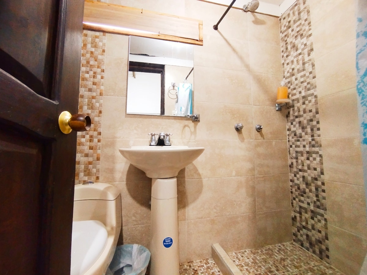 The bathroom features a modern design with neutral tilework and a glass shower. A pedestal sink with two faucets stands adjacent to a mirror. A small window allows natural light, while a soap dispenser and a candle add subtle decor.
