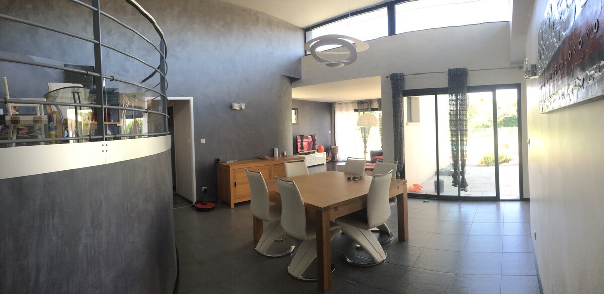 An open-plan living area is presented, featuring a large wooden dining table surrounded by white chairs. Natural light pours in through expansive glass doors, leading to an outdoor space. A modern light fixture hangs above the table, complementing the sleek gray walls.
