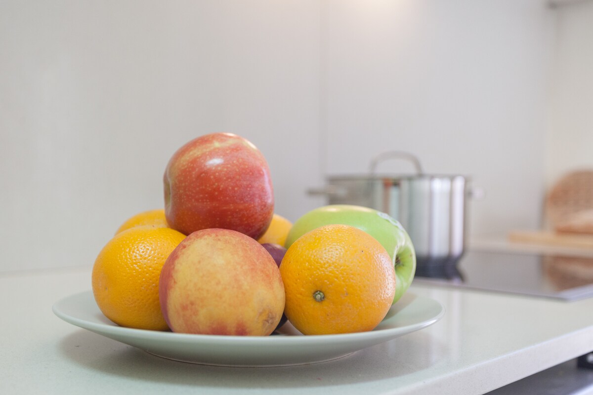 A simple plate holds a variety of fresh fruit, including red and yellow apples, oranges, and a green apple. A pot is visible in the background, suggesting a functional kitchen space.