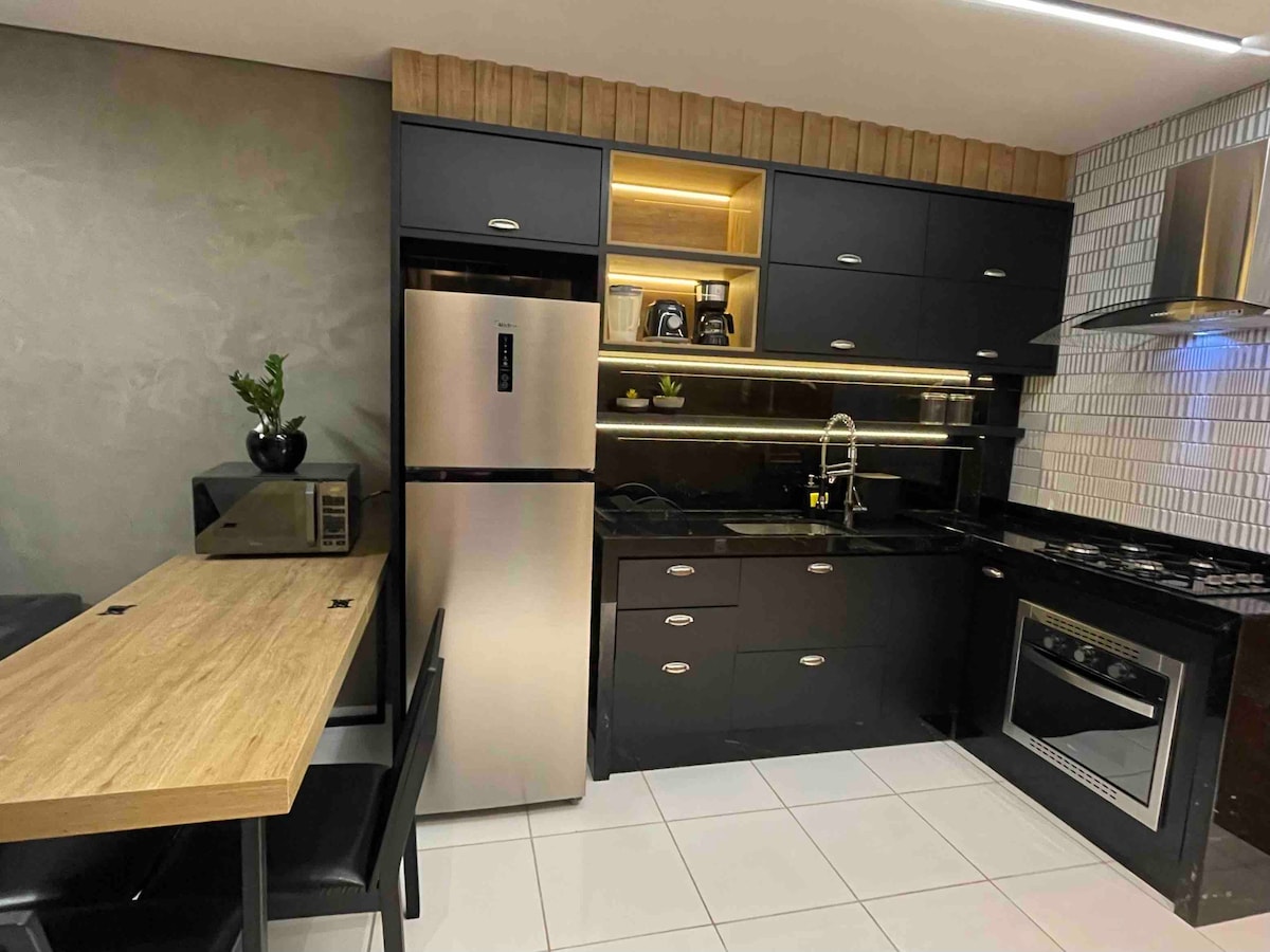 A modern kitchen is presented with sleek black cabinetry and stainless steel appliances. A refrigerator and a microwave are visible, alongside a built-in gas stove. A wooden dining table with black chairs is positioned nearby, creating a functional and practical cooking space.