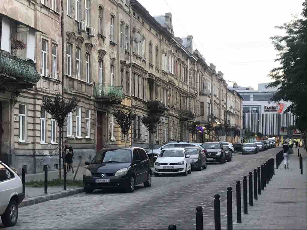A street scene showcases historic buildings with intricate architectural details. Cars are parked along the cobblestone road, while trees with hanging foliage provide a touch of greenery. The backdrop features modern structures, presenting a blend of old and new in the city center.