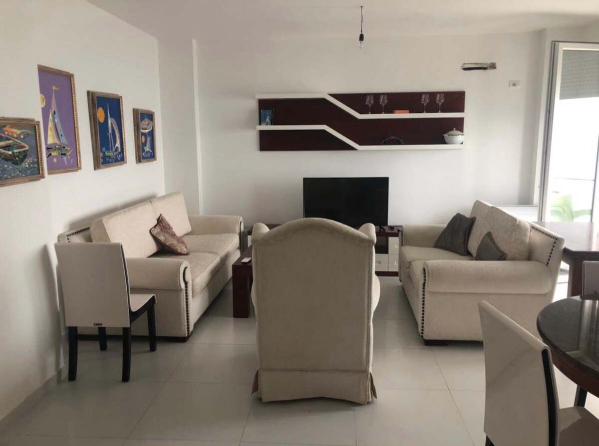 A modern living area features two light-colored sofas facing a television unit. Additional seating is provided by a cream armchair and dining chairs. Decorative artwork adorns the walls, while a sleek shelf and table enhance the contemporary design.