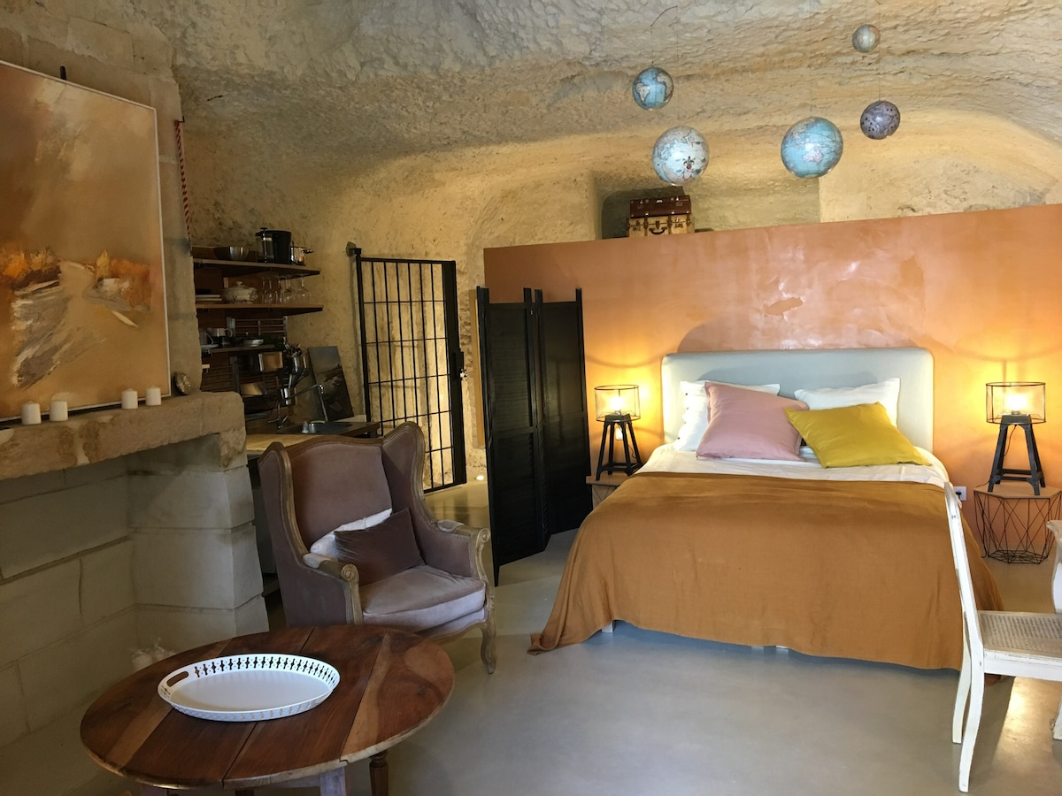 A cozy troglodyte bedroom features a spacious bed with soft linens and decorative pillows. A wooden table is positioned nearby, complemented by a comfortable armchair. Ambient lighting is provided by two side lamps, and an artwork hangs on the wall, enhancing the room's unique character.