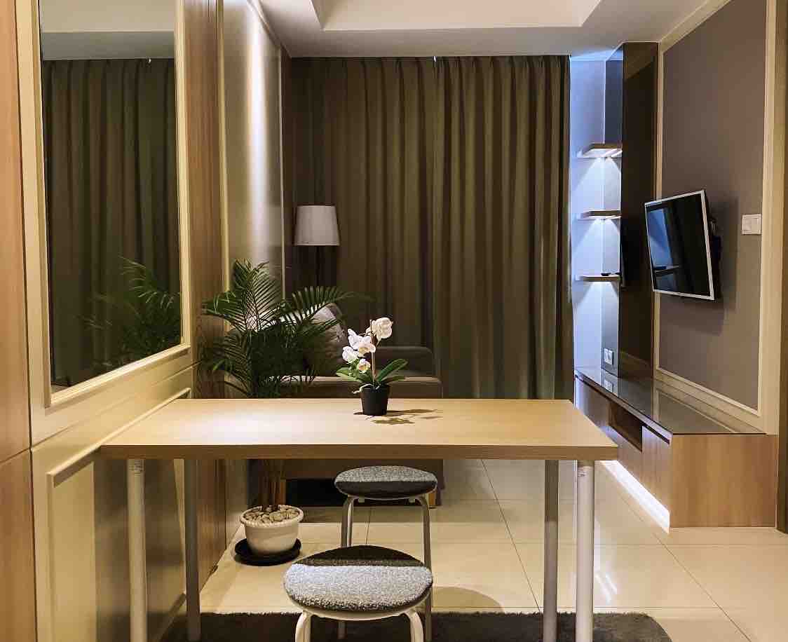 A clean and functional workspace is featured, comprising a wooden table paired with two upholstered stools. A potted orchid adds a touch of greenery. Soft lighting enhances the space, complemented by backdrop curtains and a mounted television, creating a welcoming atmosphere.