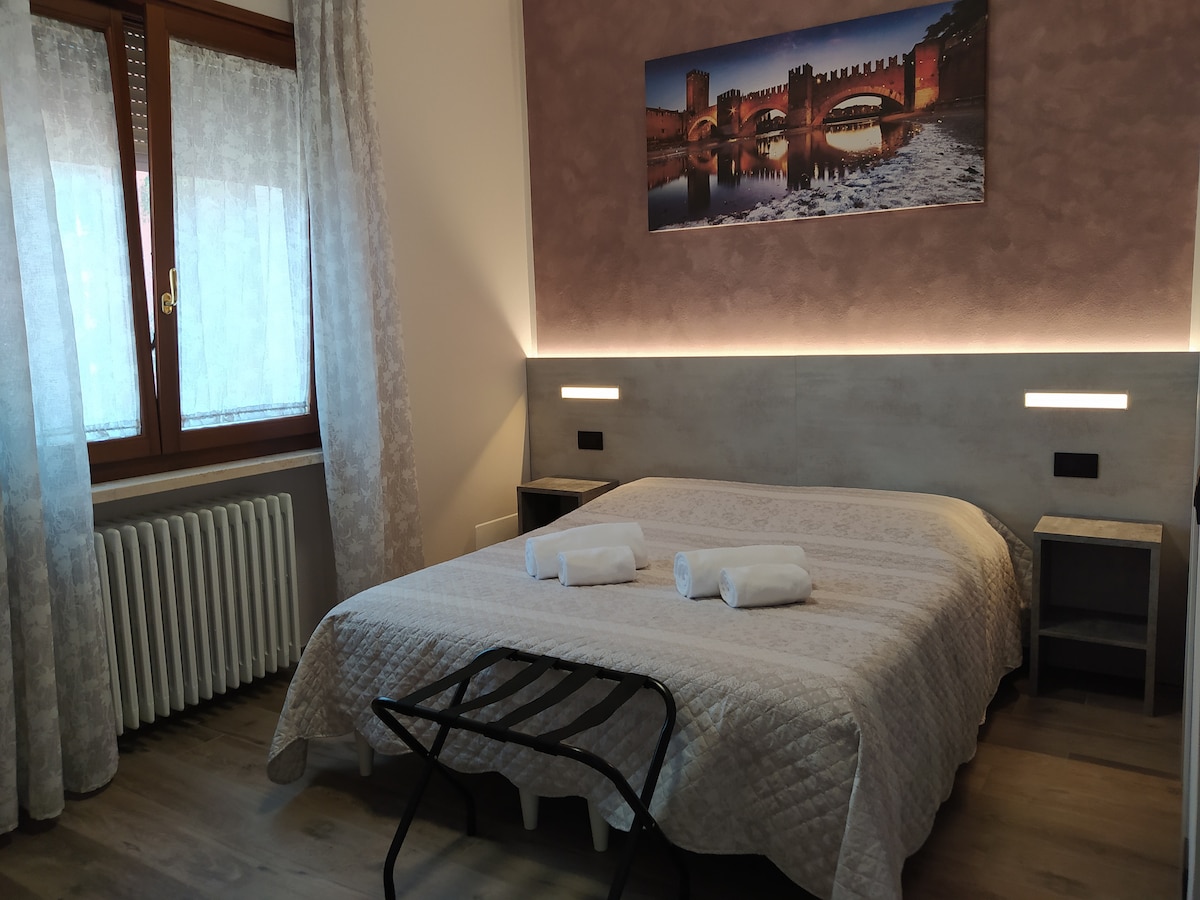 A well-appointed double bed is located against a softly colored wall, complemented by bedside lighting. Fresh white towels are neatly placed at the foot of the bed. A framed cityscape photograph adorns the wall above, offering a glimpse of the local architecture.