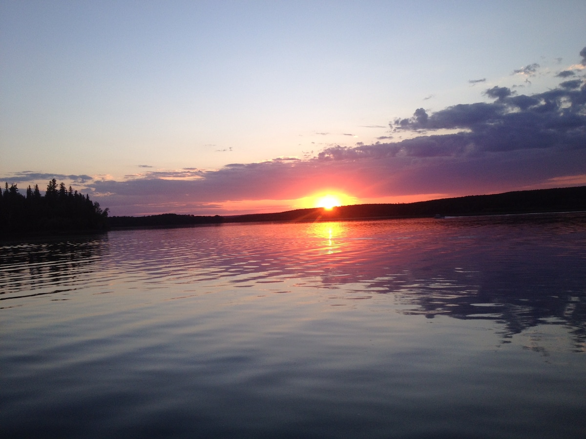 A serene lakeside view is depicted, showcasing a vibrant sunset that reflects on the calm waters. The horizon is filled with trees, creating a natural backdrop against the colorful sky as shades of orange, pink, and purple blend together.