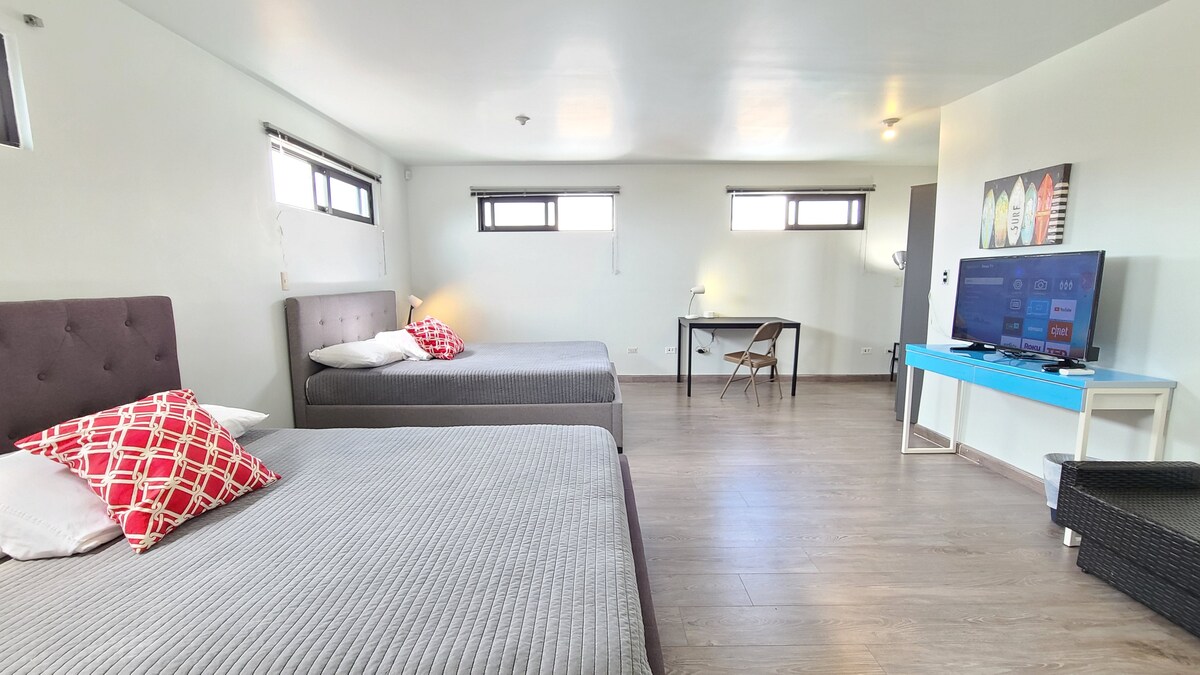A bright and spacious room is furnished with two queen beds, each adorned with simple bedding. A desk and chair are positioned against one wall, and a smart TV stands on a blue console along the opposite wall. Two windows allow natural light to fill the space.