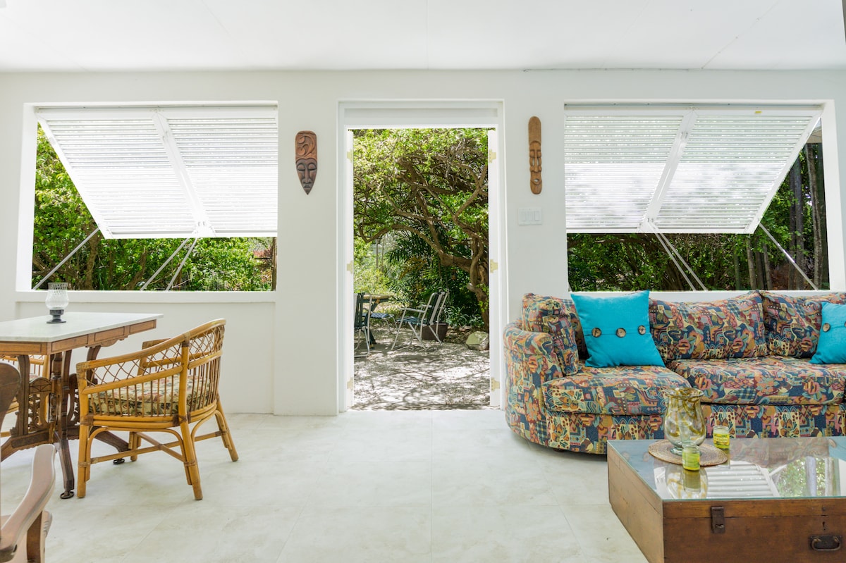 A bright living area features large windows with white blinds, allowing natural light to filter in. A patterned couch with colorful cushions is positioned next to a wooden coffee table. A dining table with woven chairs complements the space, creating a comfortable and inviting atmosphere.