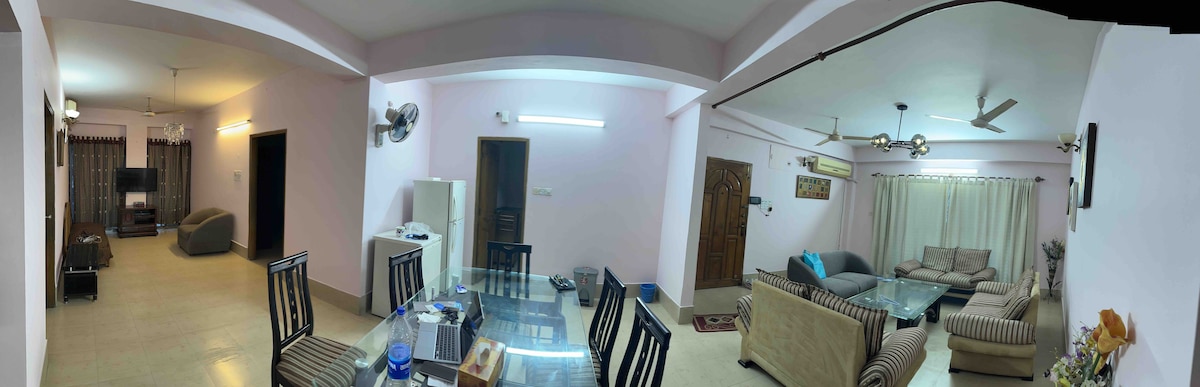 A spacious living and dining area is featured, showcasing a glass dining table surrounded by black chairs. Comfortable seating is arranged on both sides, including a light-colored sofa and a patterned armchair. Soft lighting illuminates the room, creating a relaxed ambiance.