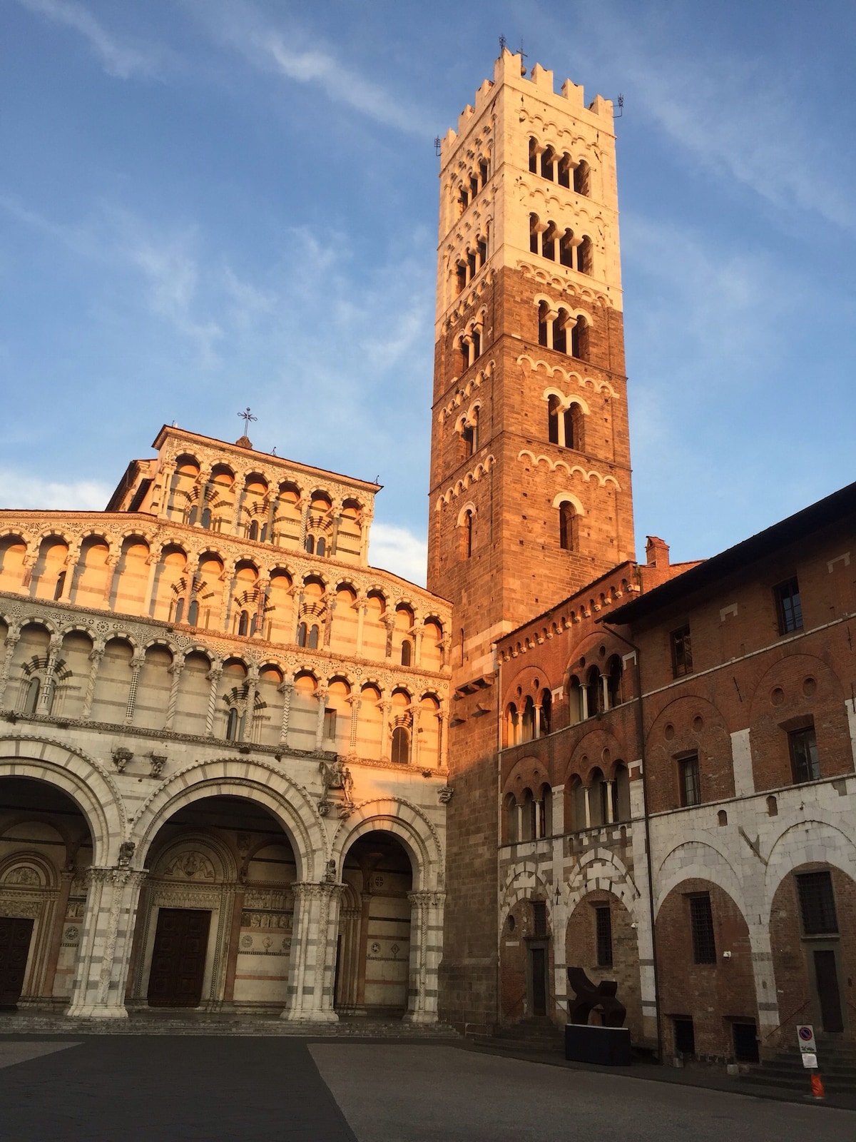 A historical building complex is captured in warm evening light, highlighting intricate architectural details. The tall bell tower, adorned with windows, stands prominently next to a beautifully designed façade featuring arches and columns. The setting reflects a blend of historic charm and cultural significance.
