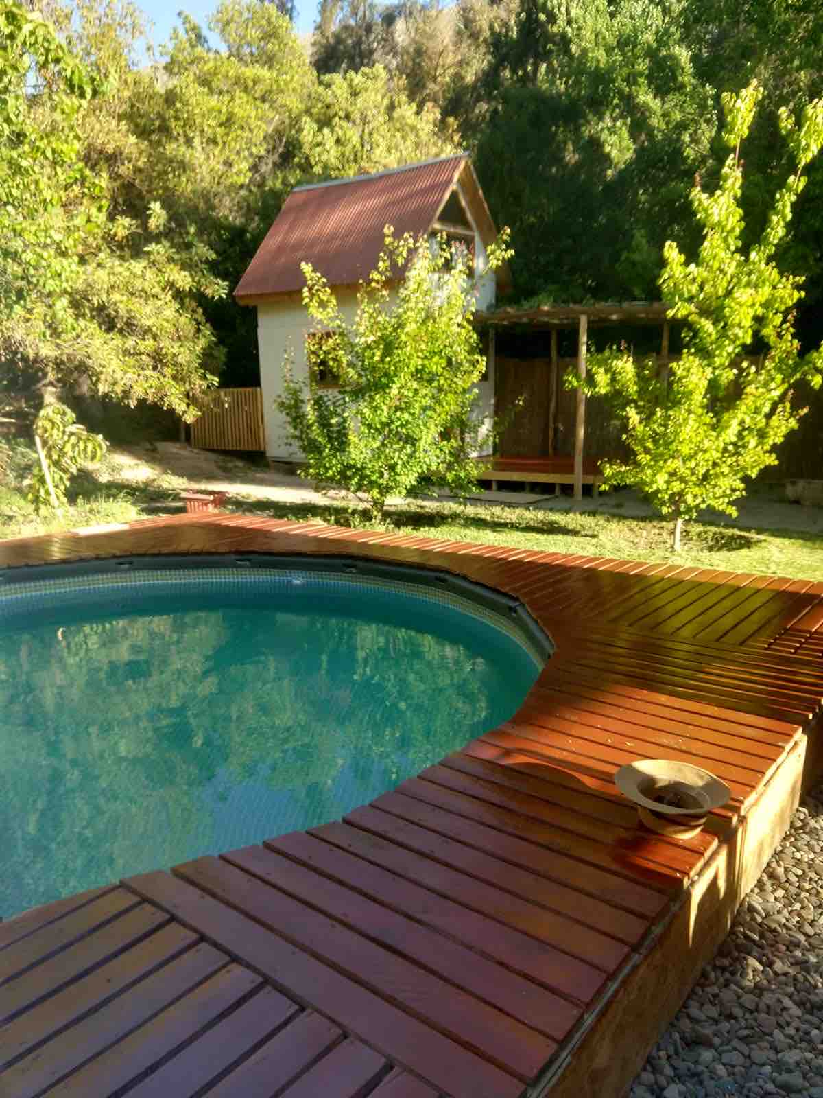 A private pool is surrounded by wooden decking, featuring a circular shape and clear water. Lush greenery, including small trees and shrubs, frames the area, creating a natural setting. A charming cottage with a red roof is visible in the background.