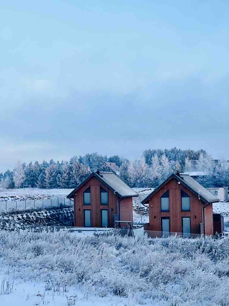 Two modern cabins are set against a snowy landscape, showcasing a mixture of stone and wooden construction. Large windows reflect the winter sky, while surrounding foliage is dusted with snow, providing a serene backdrop in the heart of nature.