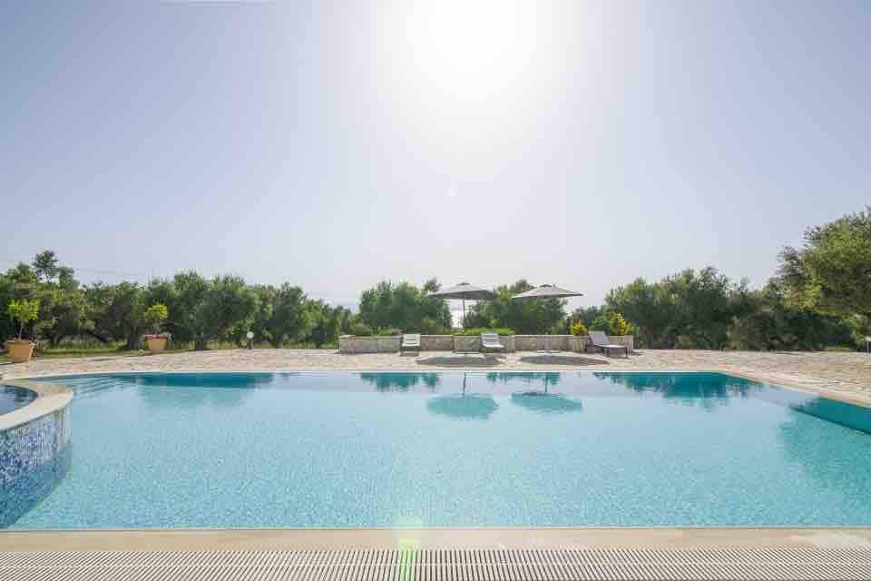 The outdoor pool is framed by lush greenery, providing a serene backdrop. Sun loungers are positioned beside the water, with large umbrellas offering shade. The sun reflects off the clear, blue water, creating a peaceful atmosphere for relaxation.
