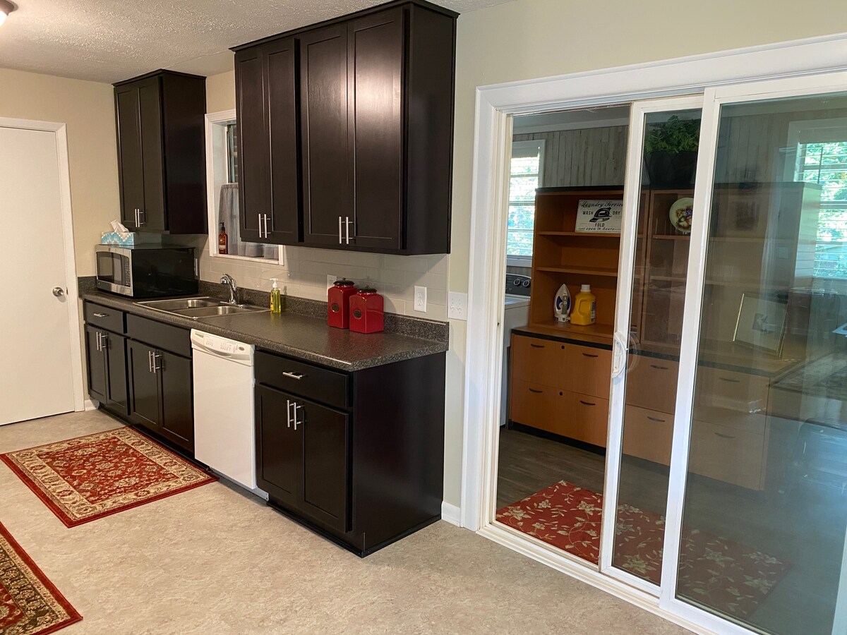 A modern kitchen features dark cabinetry and a countertop, equipped with essential appliances including a microwave and dishwasher. Natural light enters through a large window, while an adjacent room is partially visible through sliding glass doors.