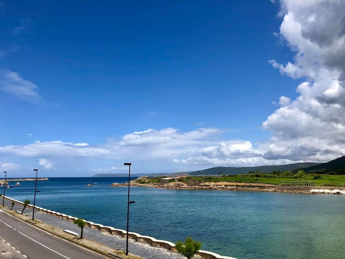 The image captures a serene coastal view, featuring calm turquoise waters bordered by a rocky shore. A clear blue sky, dotted with white clouds, stretches overhead, creating a tranquil atmosphere. A road runs parallel to the shoreline, reflecting the peaceful nature of this seaside location.