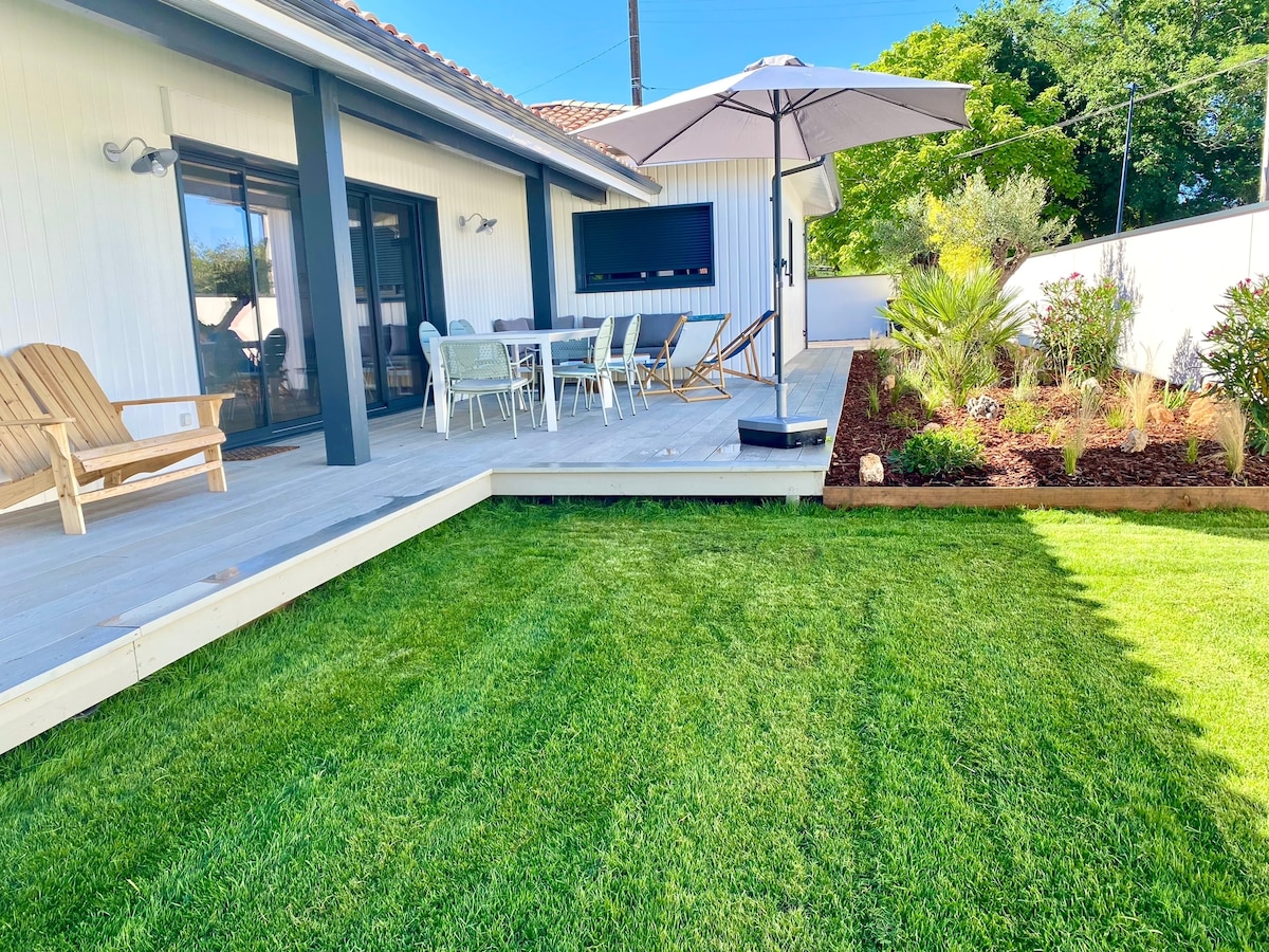 A spacious outdoor terrace is shown, featuring a dining table with several chairs under a large umbrella. A comfortable lounge chair is present, alongside well-maintained grass and garden beds filled with various plants, contributing to a serene outdoor space.