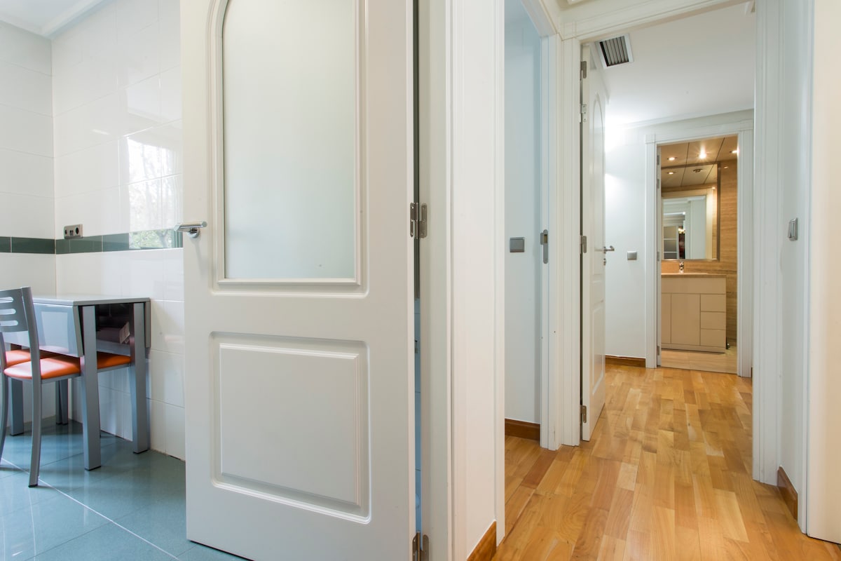A well-lit hallway is shown, featuring a wooden floor and white doors. A small dining area with a black table and two chairs with orange upholstery is visible to the left. The space is enhanced by natural light coming from the adjacent room.