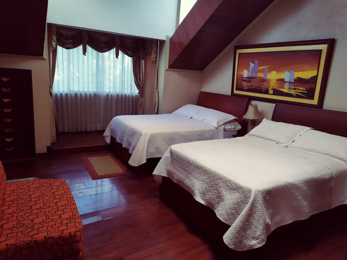 The room features two spacious beds dressed in white linens, positioned against a wall with warm wooden accents. A large window allows soft natural light to brighten the space, complemented by elegant drapery and a framed artwork above the beds. A cozy seating area is in the corner.