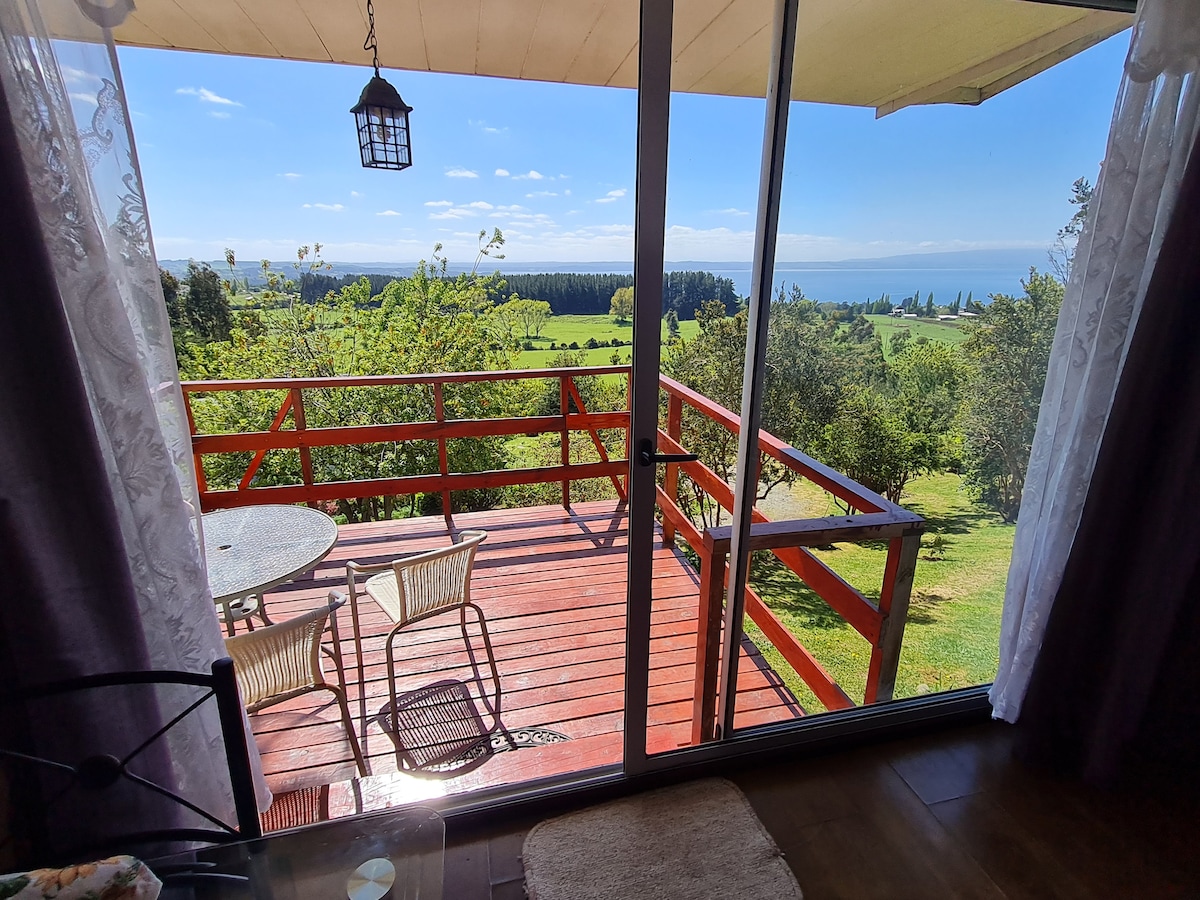 A glass slider opens to a wooden deck with a scenic view of rolling green hills and Lake Ranco. Two chairs and a small table are situated on the deck, inviting relaxation in the natural surroundings. Bright sunlight illuminates the area.
