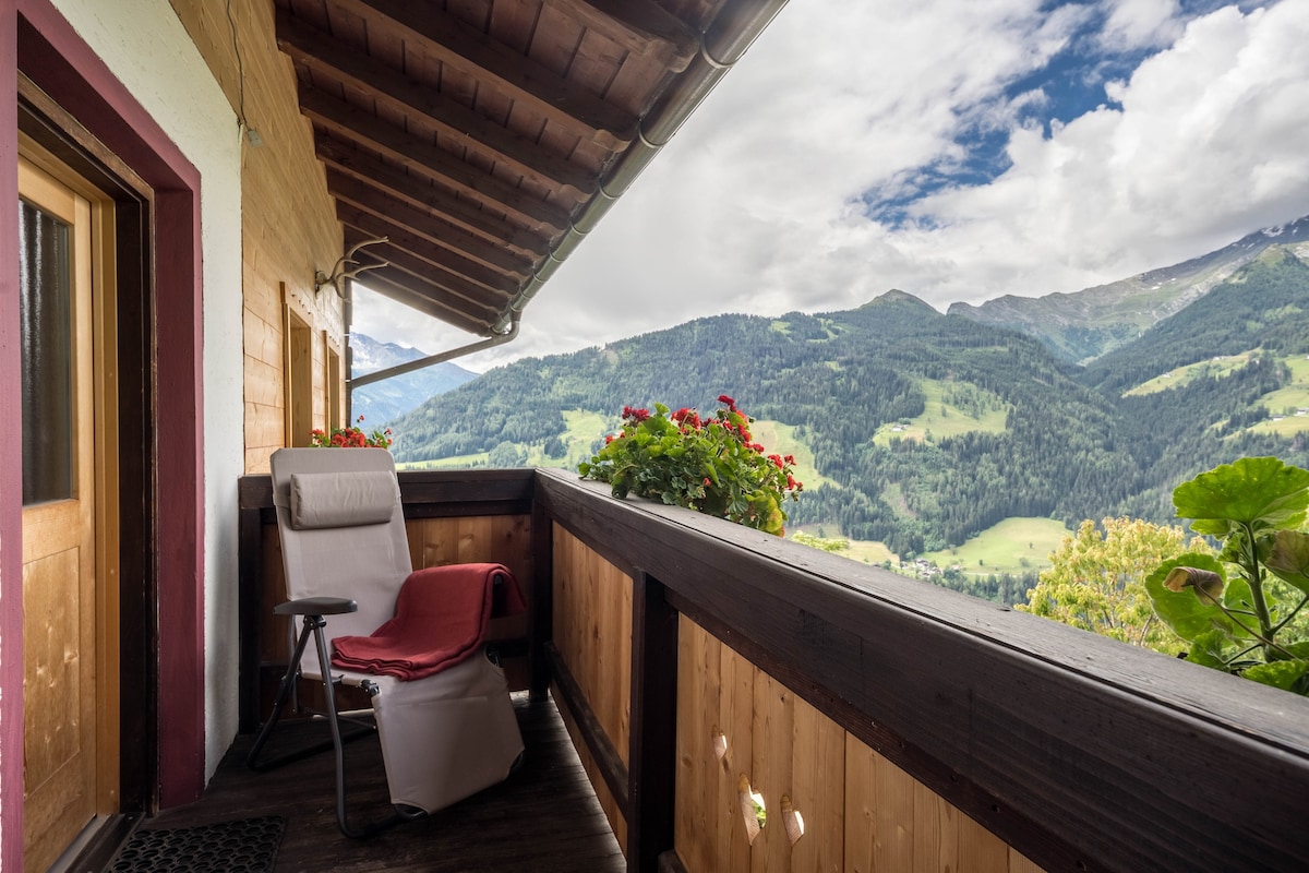 A private balcony features a comfortable chair with a red blanket draped over it, surrounded by blooming flowers. Stunning mountain views extend in the distance under a partly cloudy sky, creating a serene outdoor space for relaxation.