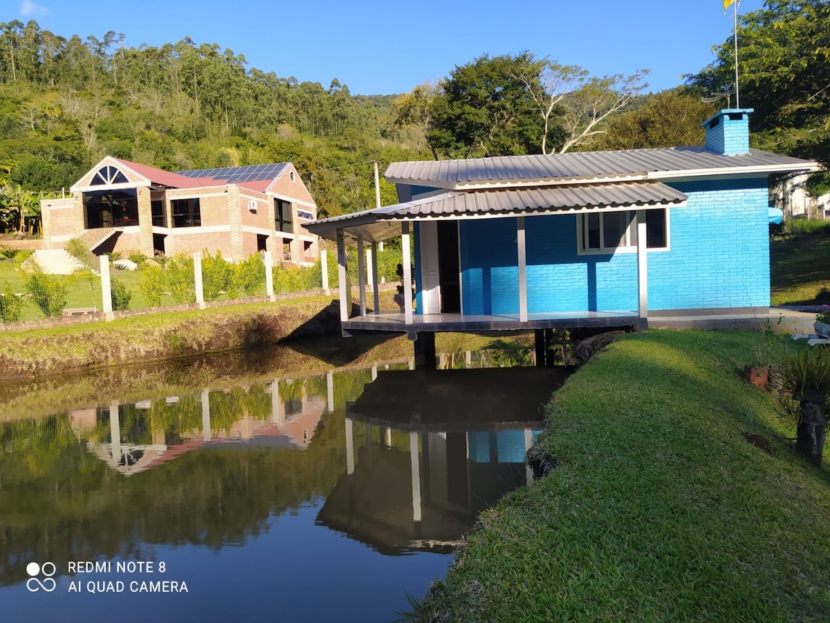 A colorful blue kiosk is situated above a calm pond, reflecting its structure in the water. Lush greenery surrounds the area, and a brick building stands in the background, adding to the tranquil setting. A gentle slope of grass leads up to the kiosk, inviting relaxation.