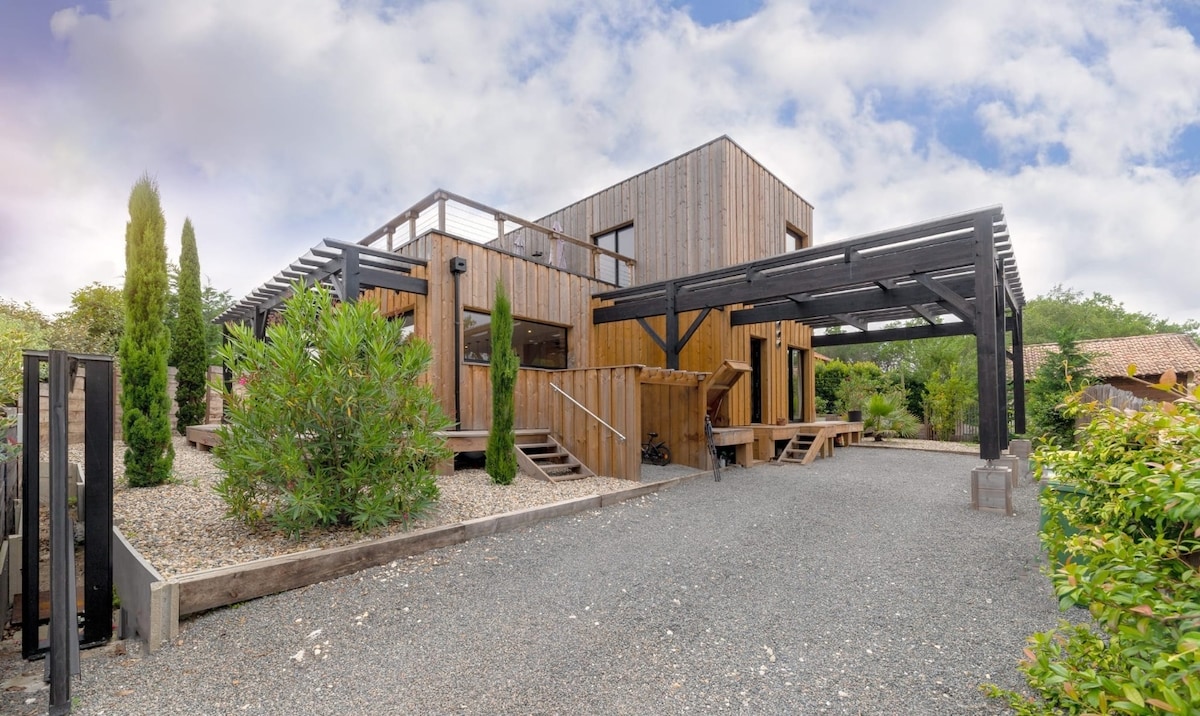 The exterior of the wooden villa is displayed, featuring a multi-level structure with a spacious gravel driveway. Lush greenery and tall trees frame the property, providing a natural atmosphere. A covered terrace and various outdoor seating areas are visible, offering places to relax.