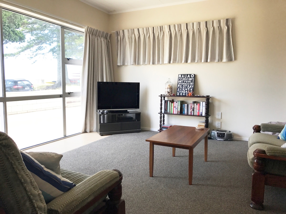 A comfortable living area features a large window with views of the outdoors. A television is positioned on a low cabinet beside a small bookshelf filled with various items. A wooden table is centered in the room, surrounded by two armchairs.
