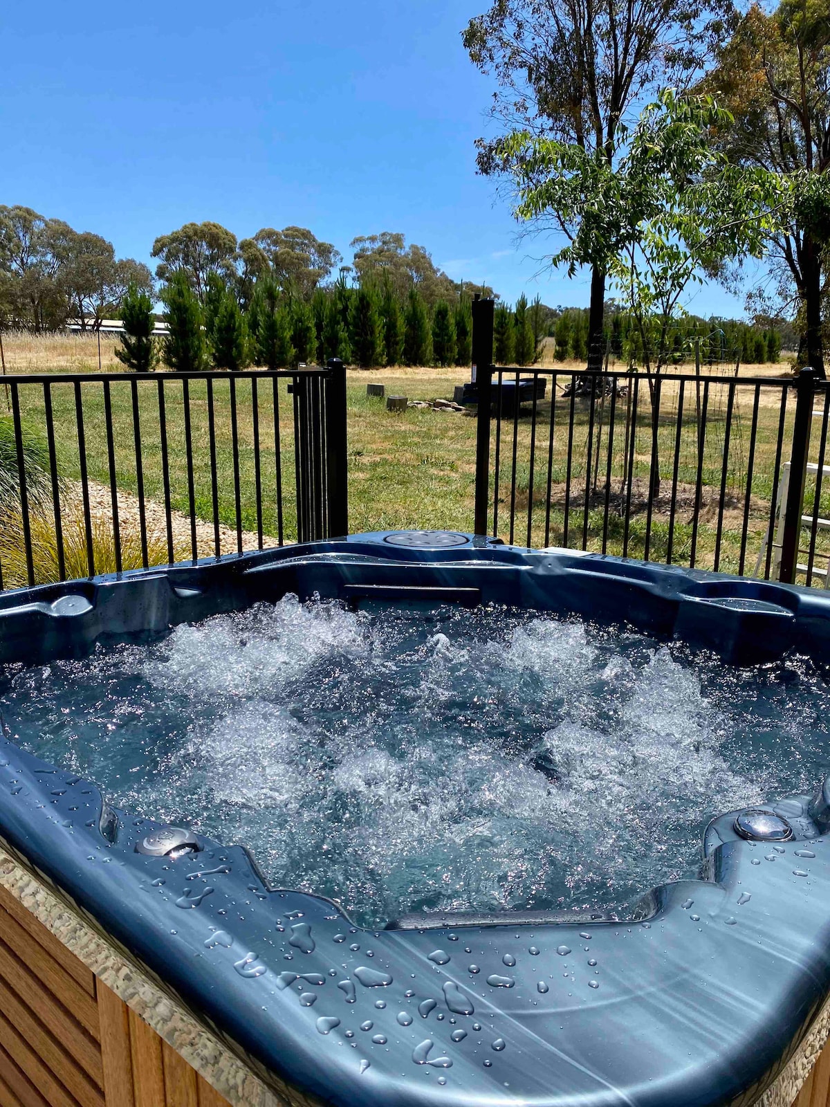 A heated outdoor spa is depicted, featuring bubbling water that creates a relaxing atmosphere. The surrounding fencing offers privacy, while a scenic view of open fields and trees is visible in the background under a clear blue sky.