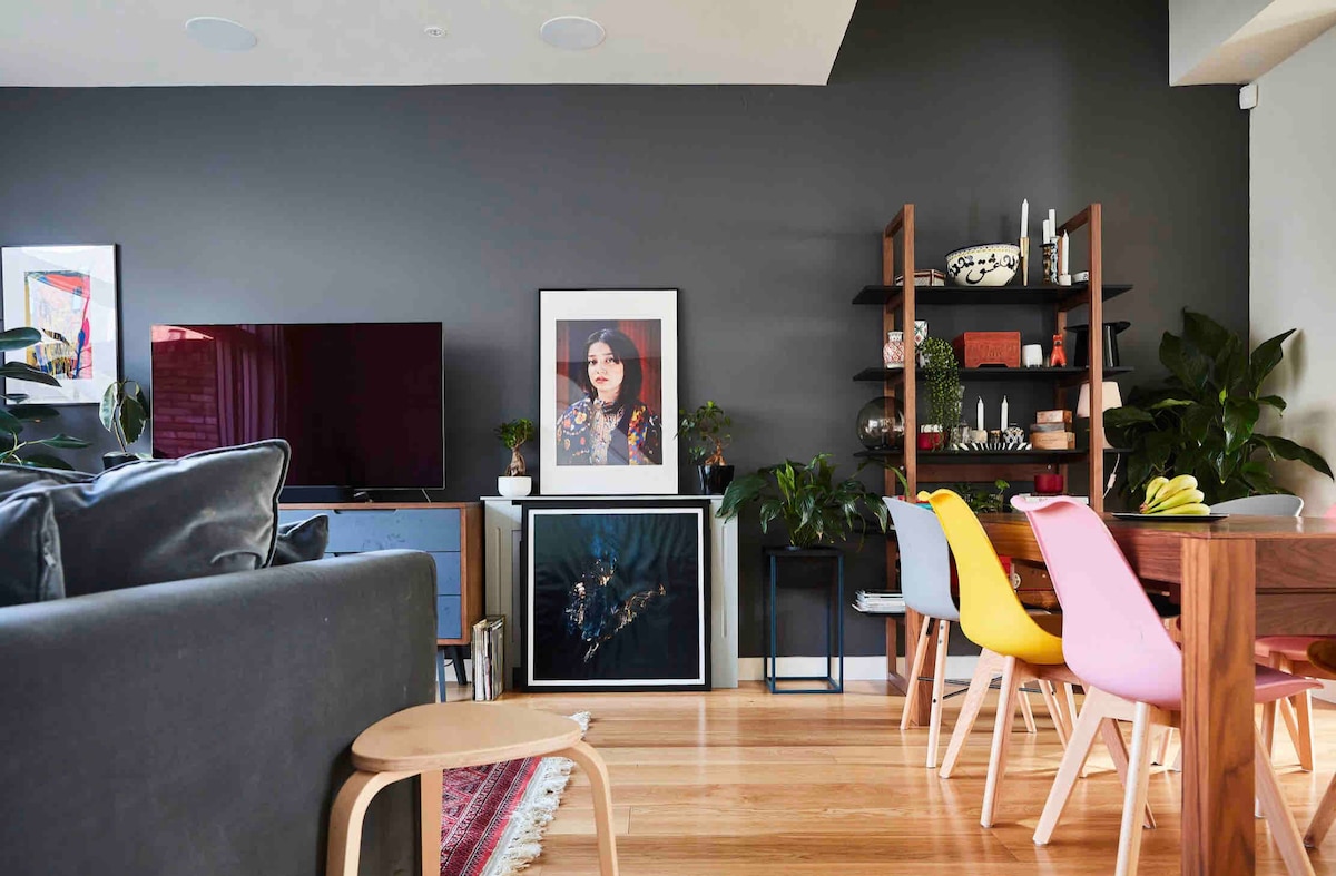 A modern living area showcases a dark accent wall, complemented by a large Smart TV and framed artwork. Wooden furniture includes a dining table with colorful chairs. Plants are arranged throughout, adding a touch of greenery to the inviting space.