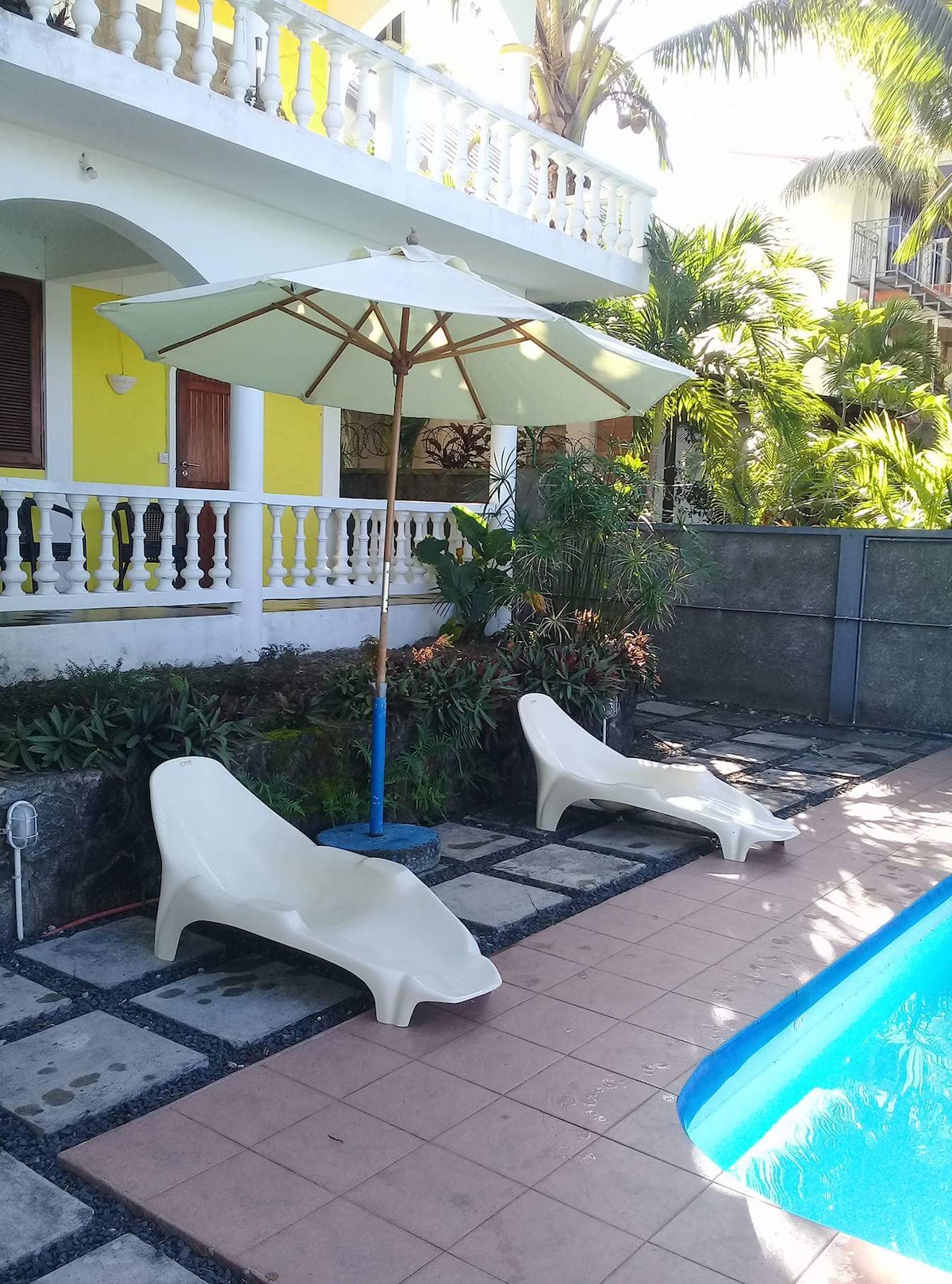 Two lounge chairs are positioned near a swimming pool, accompanied by a large umbrella providing shade. Lush greenery surrounds the area, with a tiled pathway leading to the pool. The cheerful yellow exterior of a nearby house is partially visible in the background.