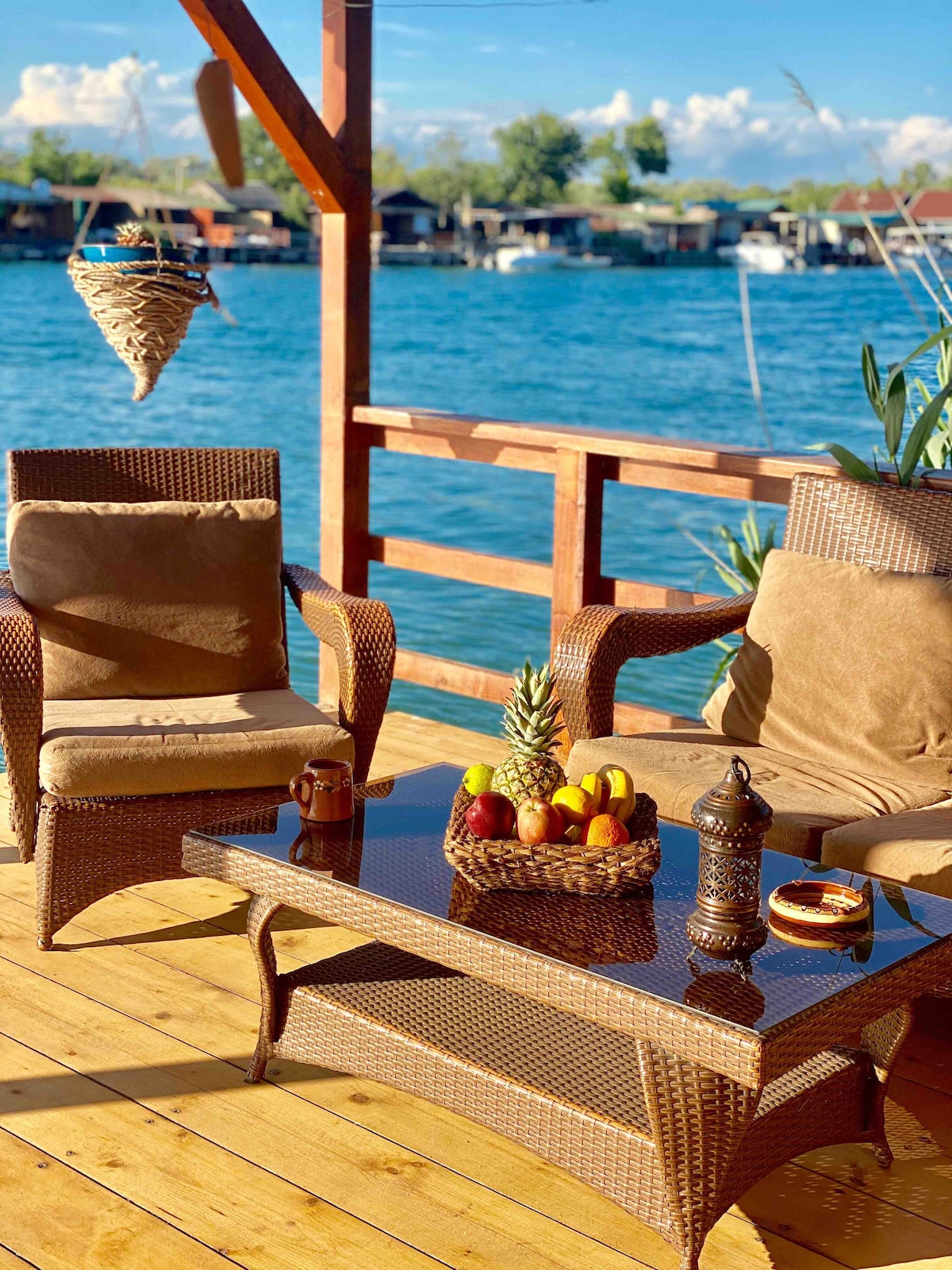 An outdoor terrace features two comfortable chairs beside a woven coffee table. A fruit basket, adorned with colorful fruits, is placed at the center. The serene water view is complemented by a gently swaying hanging decoration, enhancing the tranquil atmosphere of the space.