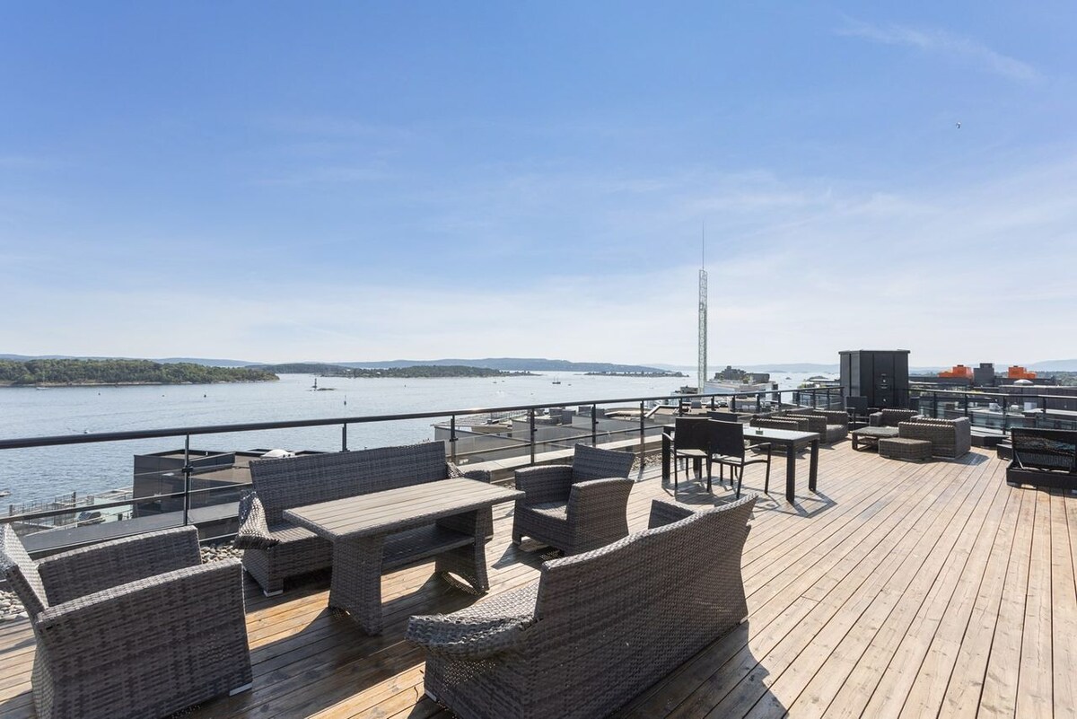 A spacious rooftop terrace offers ample seating with wicker furniture, including lounge chairs and tables. The expansive wooden deck overlooks the water, providing a clear view of the surroundings. Bright blue skies enhance the open atmosphere, making it an inviting outdoor space.
